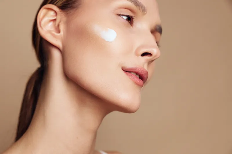 Closeup of woman with smooth skin applying white cream on cheek, natural makeup, soft lighting, skincare routine, beauty concept, beige background, healthy complexion.