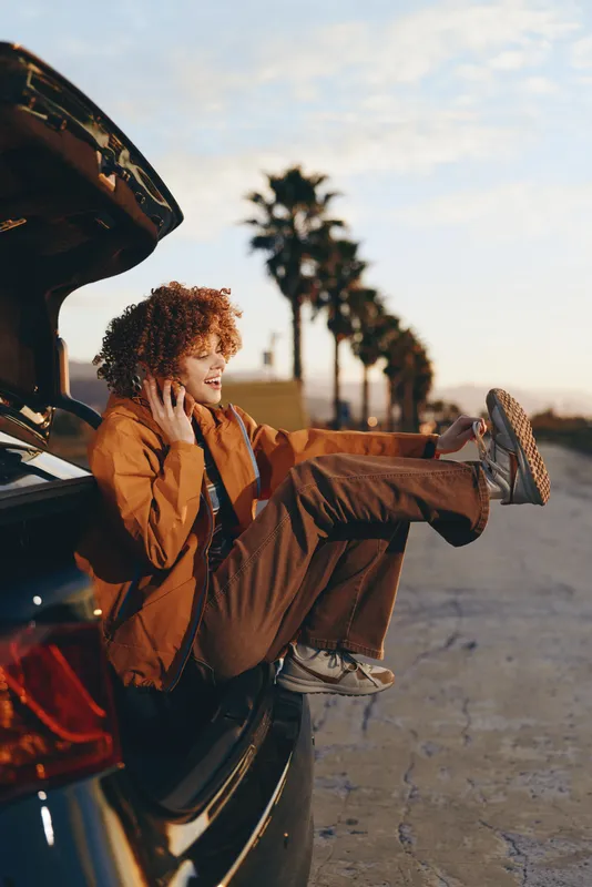 Woman with curly hair in a brown jacket by an open car door on a road at sunset, outdoor travel lifestyle, relaxed pose near vehicle, warm golden light with palm trees in background soft