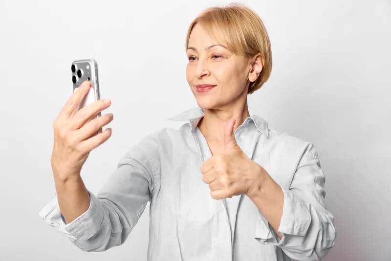 Woman smartphone selfie studio portrait pointing smiling technology communication, she holds phone for selfie, calm expression, studio lighting, neutral gray background, casual shirt