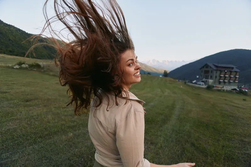 Happy woman outdoors turning with hair flying, smiling joyfully, wearing casual clothes, green meadow, mountain landscape, clear sky background