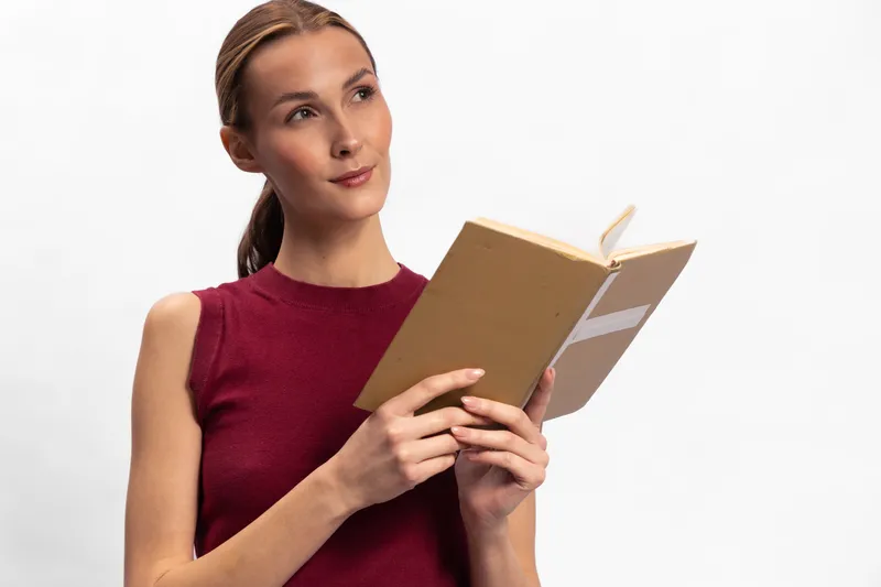 Young woman in red sleeveless top reads a book, looking thoughtfully to the side. Isolated on white background with soft lighting. Concept of education, learning, and contemplation.