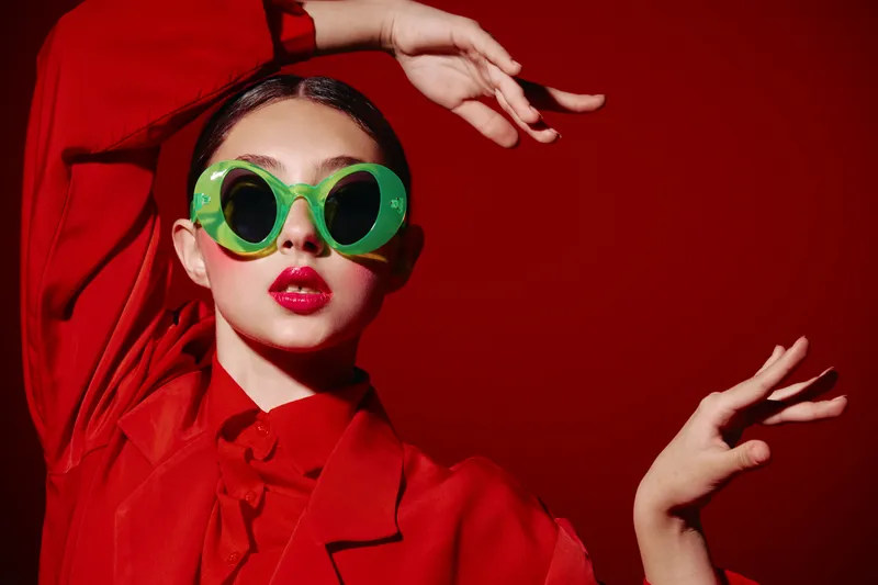 Young woman with glossy red lips wearing stylish oversized green sunglasses and red outfit poses expressively on a vibrant red background, fashion creative beauty concept.