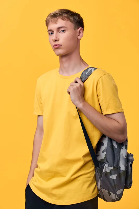 Student with a camouflage backpack and yellow t shirt stands against a solid backdrop, conveying casual education, daily learning, and youth energy for school and campus scenes