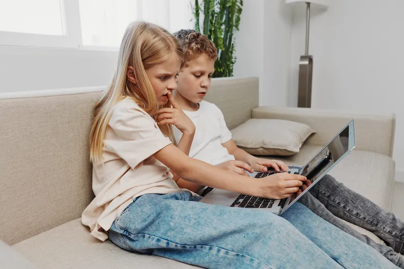 Two focused children sitting on a beige couch using a laptop together in a bright modern room. Concept of technology, learning, collaboration, and childhood curiosity.
