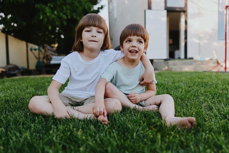 Two happy boys sitting together on a green lawn, enjoying the warm sun and the outdoors. Their cheerful smiles reflect the joy of childhood moments shared with siblings.