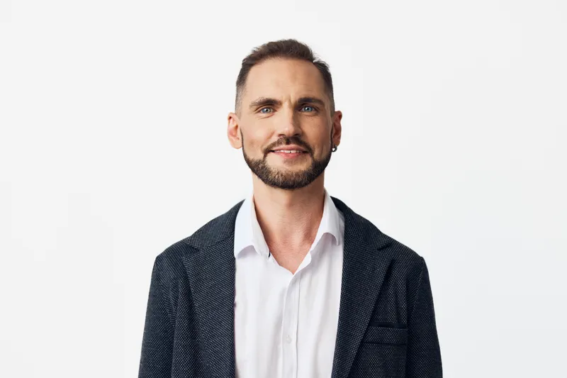 A professional man in a dark blazer and white shirt stands against a clean, isolated colored background, exuding confidence and calm. His relaxed smile signals focus and leadership.