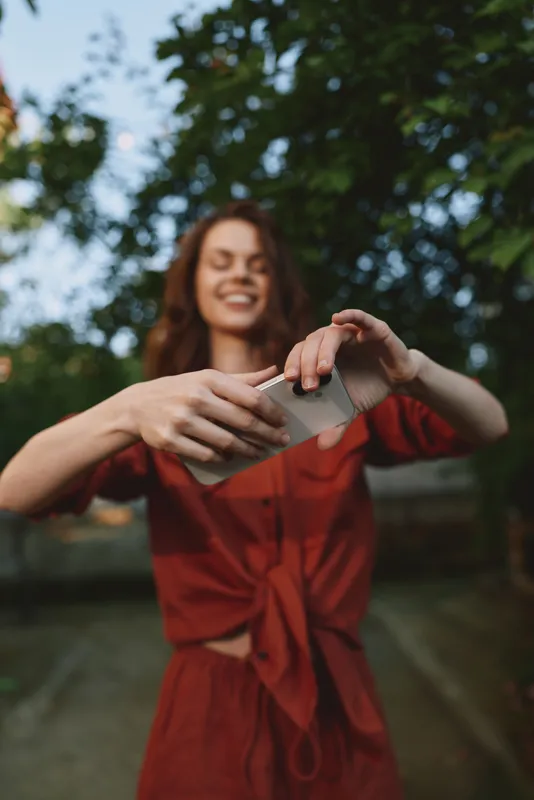 Smiling woman in a red dress outdoors, capturing a playful selfie. Wide shot with trees and sky, emphasizing carefree lifestyle, outdoor joy, and natural light. Soft daylight warmth