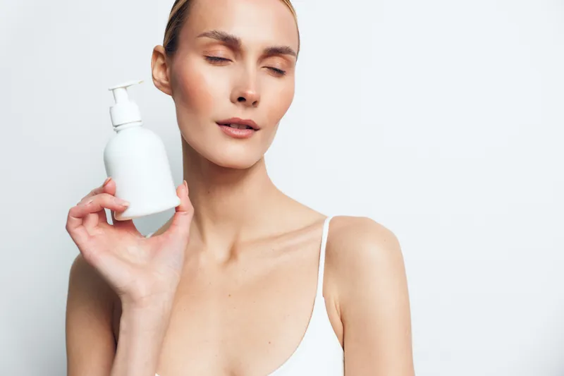 Calm woman with closed eyes holds a white bottle, applying skincare lotion to her face. Soft studio lighting on a bright background, highlighting selfcare, beauty, and wellness.