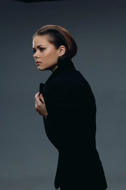Serious woman in black coat looking sideways with focused expression against dark gray background