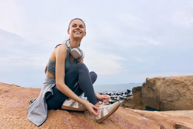 Young woman in athletic wear tying shoes on rocky beach, smiling and enjoying outdoor exercise, healthy lifestyle concept with sea in background