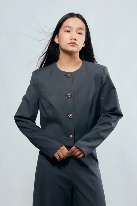 Young woman with long hair wearing a stylish gray suit with buttons standing against a light background. Confident pose with hands clasped in front of body in fashion portrait.