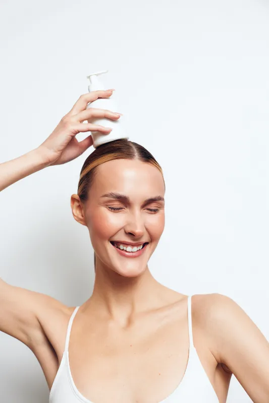 Young woman with brunette hair smiles with closed eyes while holding a white lotion bottle above her head against a plain white background in a bright skincare concept.