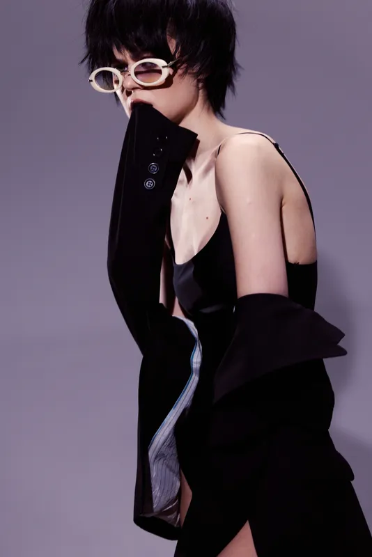 Stylish woman wearing black outfit and white sunglasses posing against gray background with confident expression.