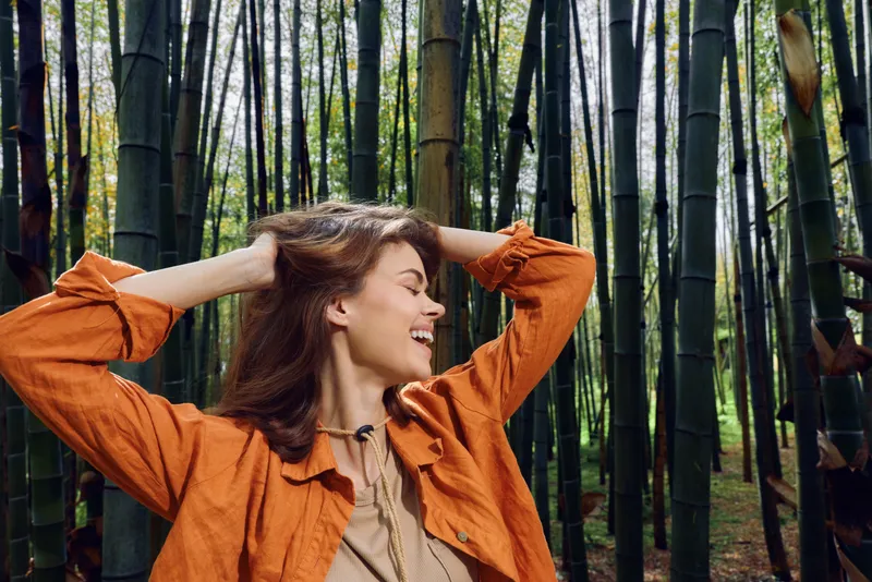 Woman bamboo forest smile nature portrait: joyful young woman in orange jacket leans back with hands in her hair among tall green bamboo stalks, enjoying peaceful outdoor relaxation.