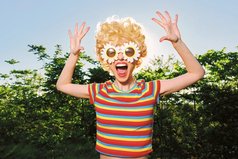 A joyful child outdoors in a striped retro outfit with oversized floral sunglasses, expressing carefree summer energy and playful fashion in bright sunlight, with a vivid garden backdrop.