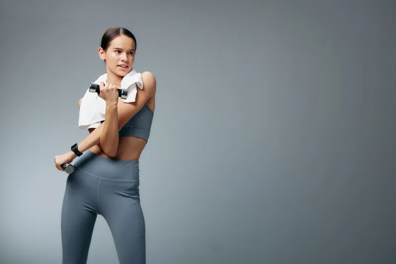 Female fitness model lifting dumbbells in athletic wear against a neutral backdrop, showcasing strength, posture and focus during a studio workout session.