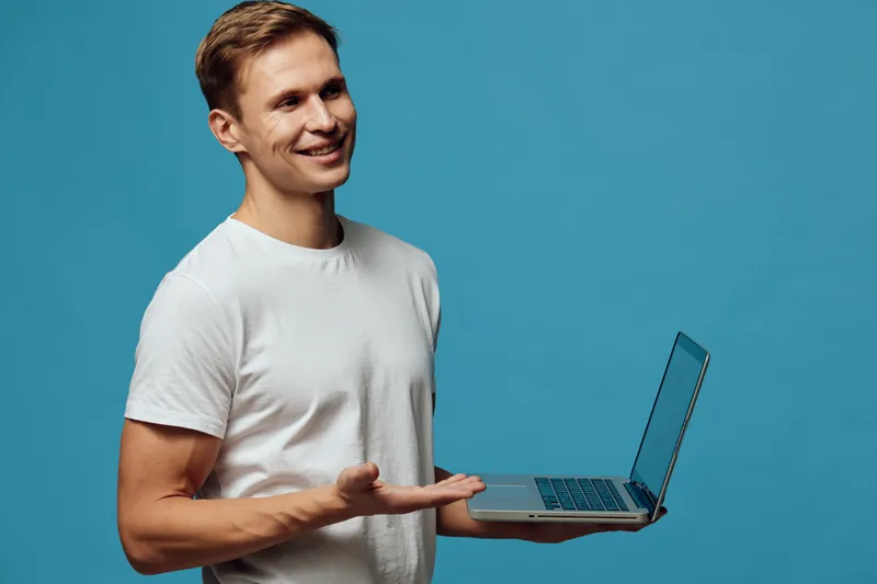Smiling man holding a laptop computer with a blue background, casual white t-shirt, positive expression, modern technology, remote work, digital device, studio shot.