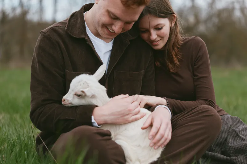 Young couple sits on grass holding a white baby goat together while enjoying a peaceful moment in nature with relaxed expressions and casual brown clothing.