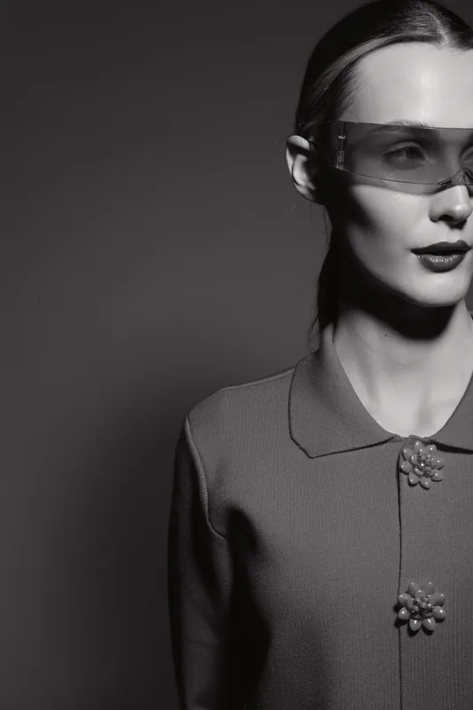 Black and white portrait of a Caucasian woman in vintage style with sunglasses and textured jacket on dark background.