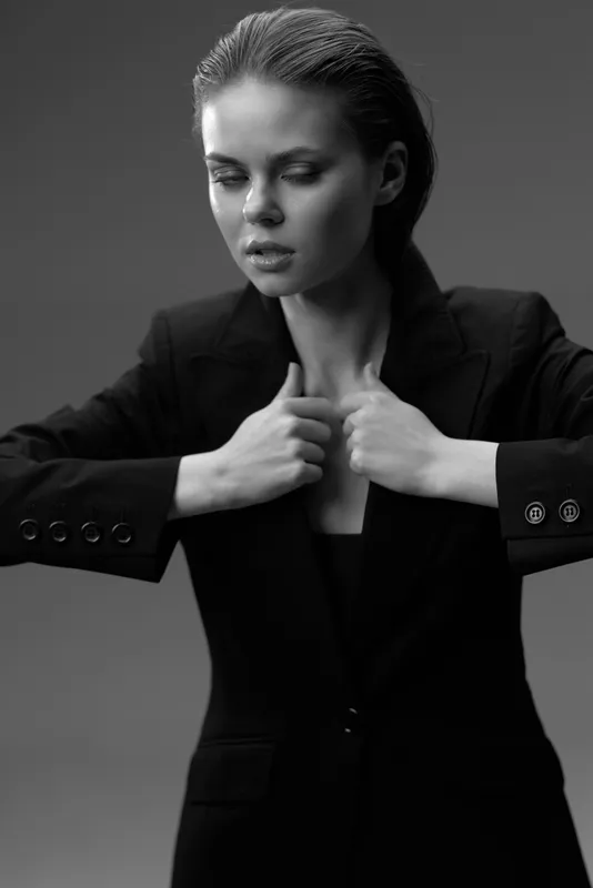 Black and white portrait of a young woman adjusting her jacket with a serious expression and sleek hairstyle.
