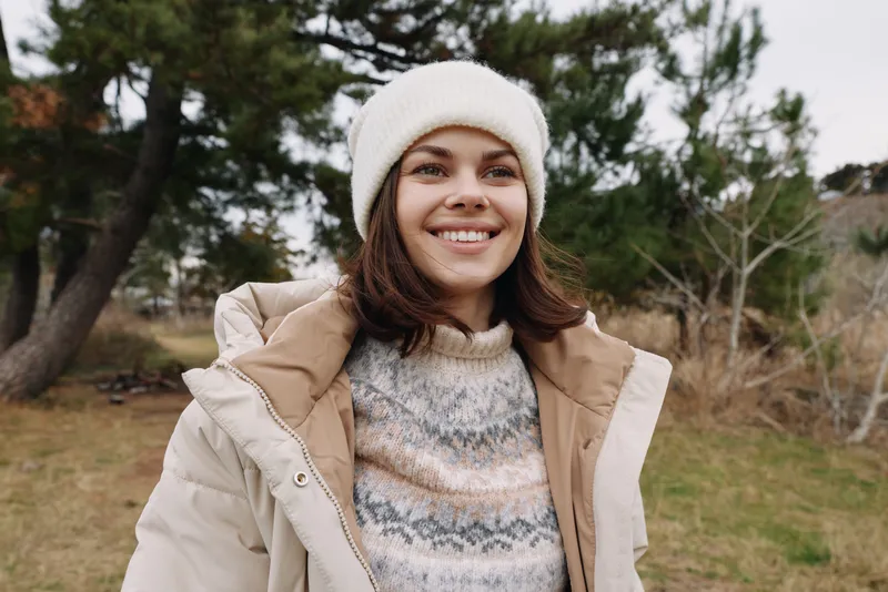 Portrait of a smiling woman outdoors in winter, wearing a light coat and knit beanie, with trees and open grass in the background.