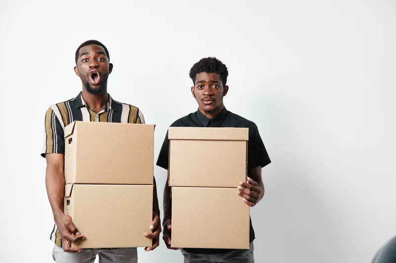 Two young black men holding cardboard boxes with surprised and neutral expressions on white background. African ethnicity, dark skin, casual clothing and moving concept.