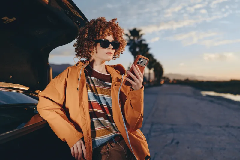 Woman lifestyle outdoors at sunset, curly haired young woman in orange jacket holds a smartphone while leaning beside an open car door, warm evening light, street background.