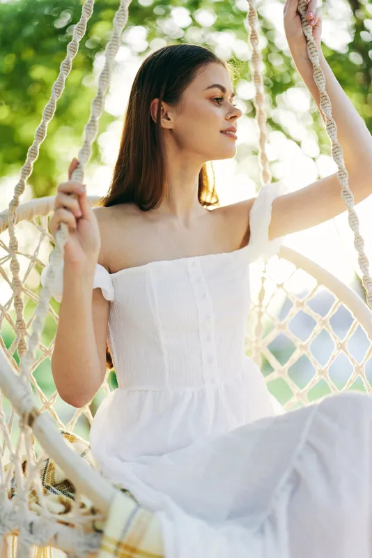 Young woman in a white off shoulder dress relaxing on a swing in a sunny outdoor setting, showcasing a tranquil summer vibe and natural beauty