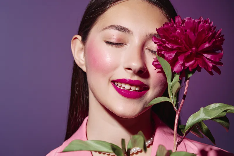 Young woman with fashion makeup and vibrant fuchsia lips poses with closed eyes near a matching pink flower. Artistic portrait with bold colors and creative beauty concept.