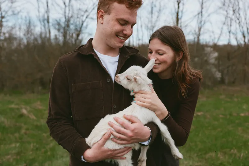 An outdoors couple warmly holds a goat kid, smiling as they cradle the animal together. This lifestyle scene emphasizes affection, bond, care, and simple farm living in bright daylight.