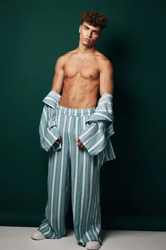 Young man with curly hair posing shirtless wearing oversized striped pants and matching open shirt against dark background in studio. Fashion, casual style and confident expression.