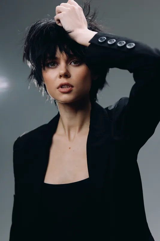 Young woman with short black hair and black outfit posing thoughtfully against dark background.