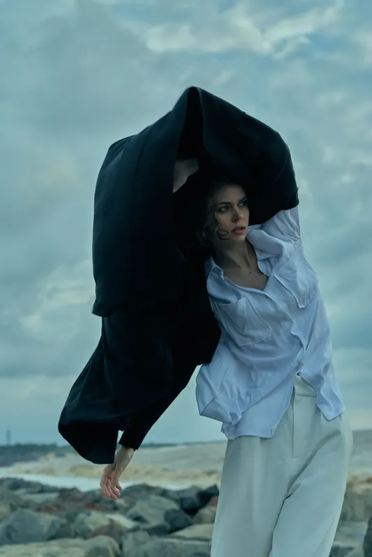 Cloak billows over a wind swept beach as a woman in a white shirt and light trousers strides forward, creating a dramatic cinematic scene with moody skies