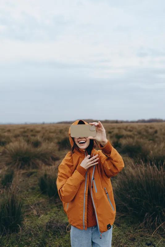 Woman lifestyle outdoors in grassy field, wearing autumn jacket and hat, holding sunglasses with both hands, cloudy sky, natural light, calm playful mood
