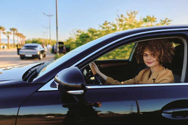 woman driving a car outdoors, cheerful smile, sunlit moment, relaxed posture, retro film color tones, casual fashion, urban street backdrop, natural lifestyle scene