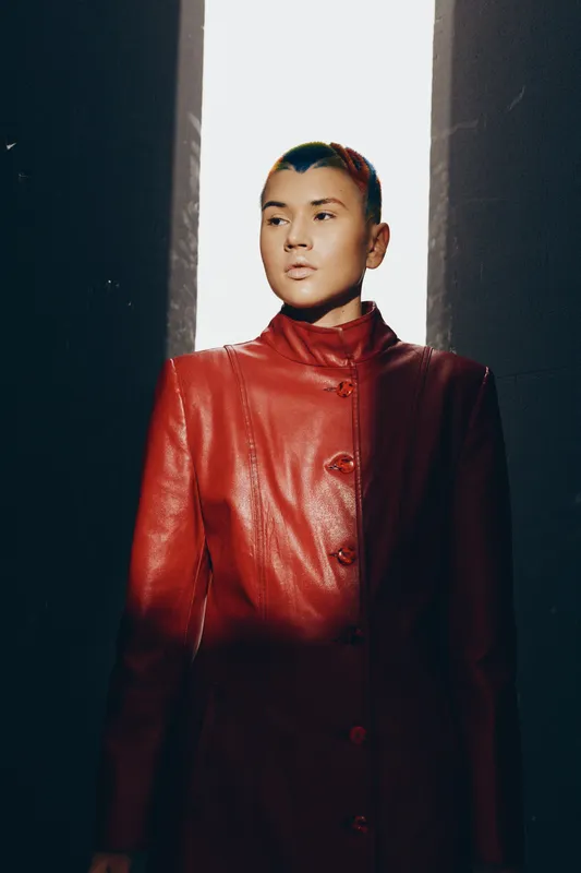 Woman wearing a stylish red leather coat stands in dramatic lighting between dark walls, showcasing bold fashion and modern urban attitude in a high-contrast setting.