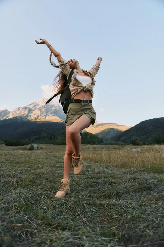 A young woman with a backpack leaps across a grassy meadow beneath a bright blue sky, radiating freedom and energy during an outdoor adventure.