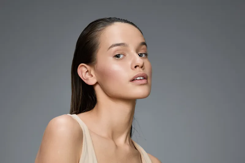 Confident young woman with natural makeup and wet hair against a neutral gray background, showcasing beauty and elegance in a fashion photography style