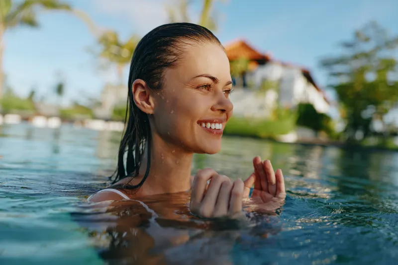 Woman enjoying sunlit outdoors, waist-deep in clear water, smiling with eyes closed, relaxed posture, soft breeze, natural colors, tropical backdrop, serene lifestyle moment