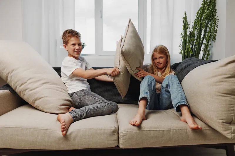 Two happy children having a pillow fight on a beige sofa in a bright living room with natural light from a window. Concept of childhood fun, playfulness, and sibling bonding.