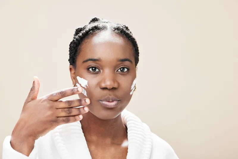 Young Black woman applying skincare product on her face, emphasizing a healthy skincare routine and glowing skin in a soft neutral backdrop