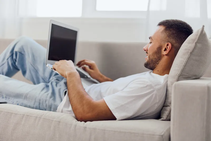 Relaxed man working on laptop comfortably seated on a couch, showcasing modern home office vibes with a stylish interior design in a light atmosphere.