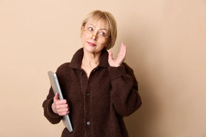 Senior woman with glasses holds smartphone in indoor beige portrait, making a friendly peace gesture with her hand, smiling calmly, wearing a dark coat, copy space for text