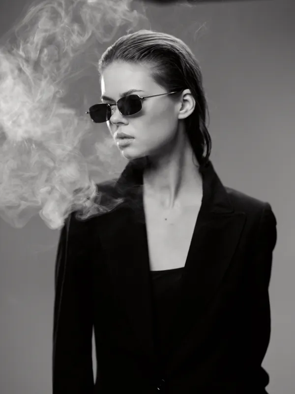 Black and white portrait of a stylish woman wearing sunglasses and a black blazer with smoke around her.