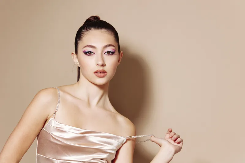 Young model in a shiny satin dress poses elegantly against beige background. Fashion beauty styling with feminine makeup and creative posture. Concept for luxury and glamour.