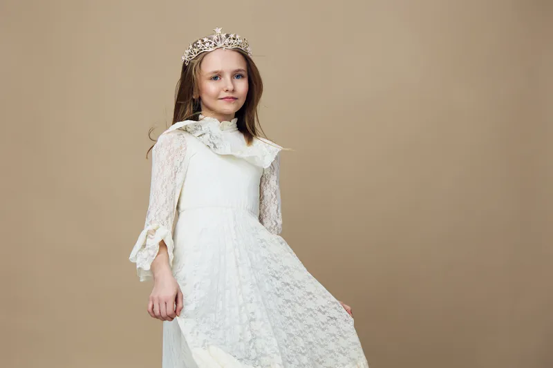 Portrait of a young girl in a white lace dress with a crown, conveying innocence and joy against a neutral background, ideal for celebrating childhood and special occasions
