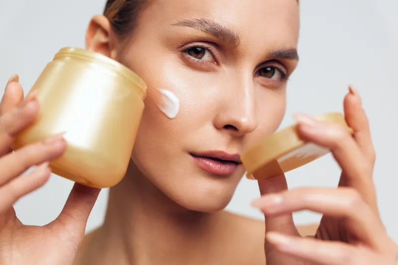 Young woman with brunette hair applies cosmetic cream from jars in a close up studio shot, capturing a skincare moment and calm self care on a neutral background with soft lighting.