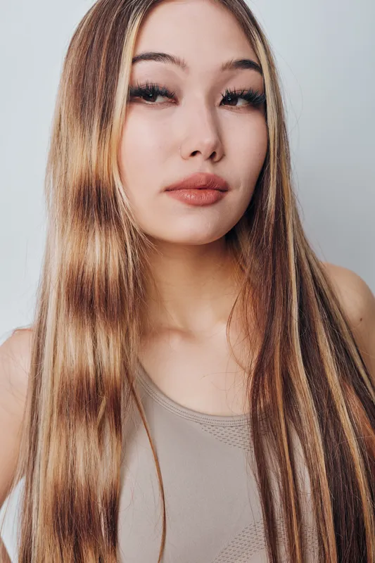 Young woman with long straight hair and natural makeup posing in casual tank top against light background, demonstrating beauty and confident expression.