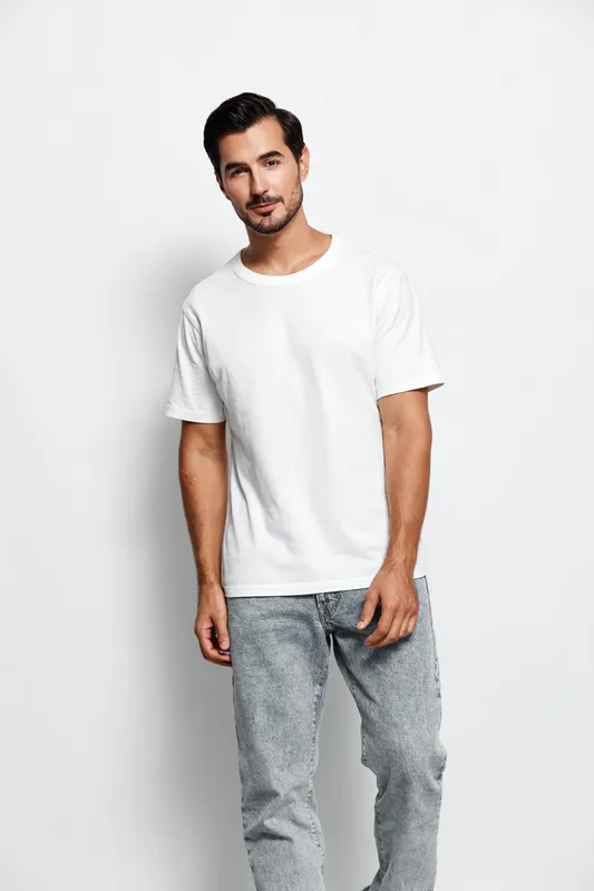 Handsome young man wearing a white t-shirt and gray jeans poses confidently for the camera while smiling. Isolated on plain solid white background studio portrait.