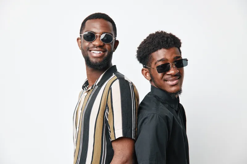 Two African American men with dark skin wearing sunglasses and casual shirts standing back to back smiling on white background. Young friends with confident expressions and stylish looks.